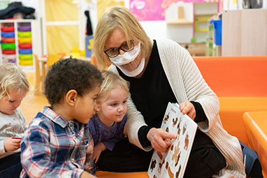 une auxiliaire de puériculture porte un masque transparent et montre un livre à 2 jeunes enfants